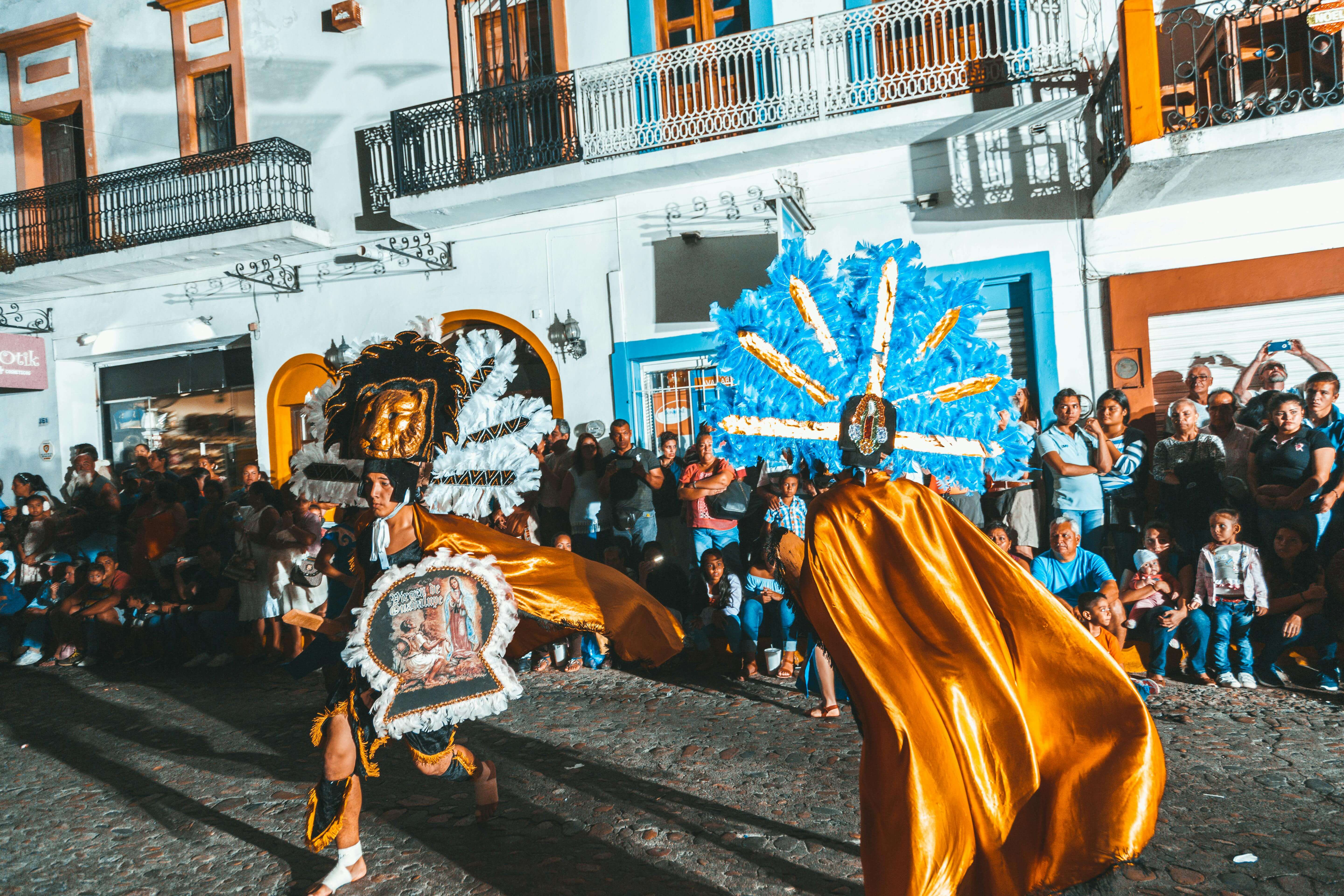 Two dancers in elaborate traditional feathered headdresses and golden costumes performing in a street parade with a crowd watching.
